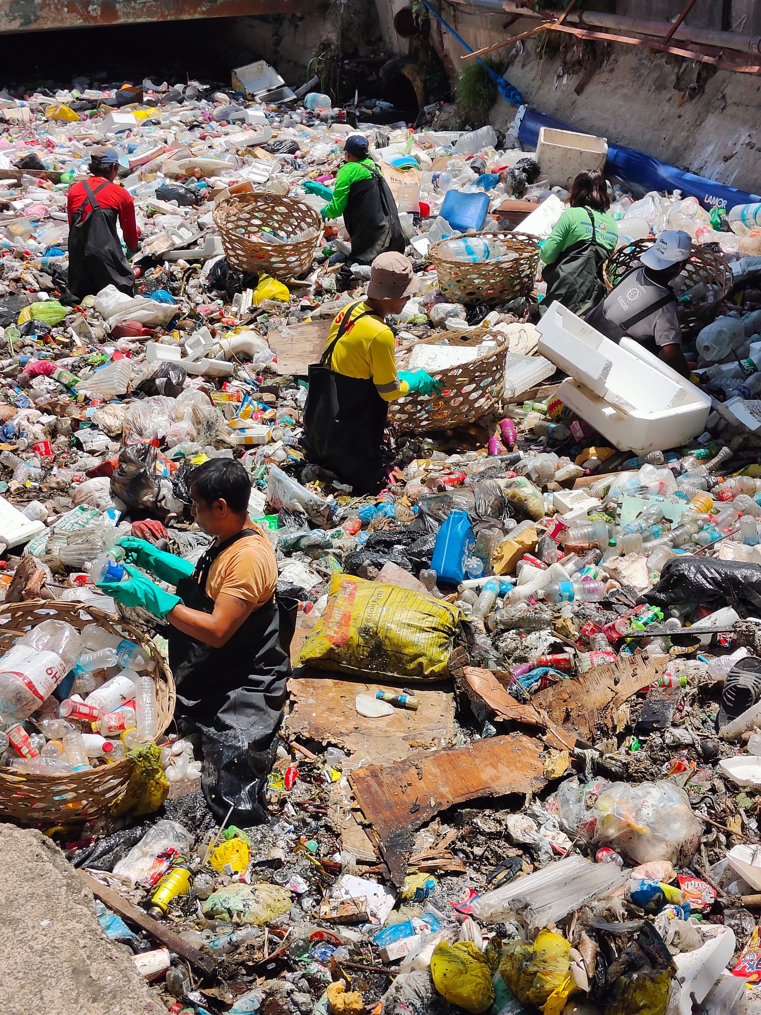 Workers with woven baskets collecting plastic waste from a river channel